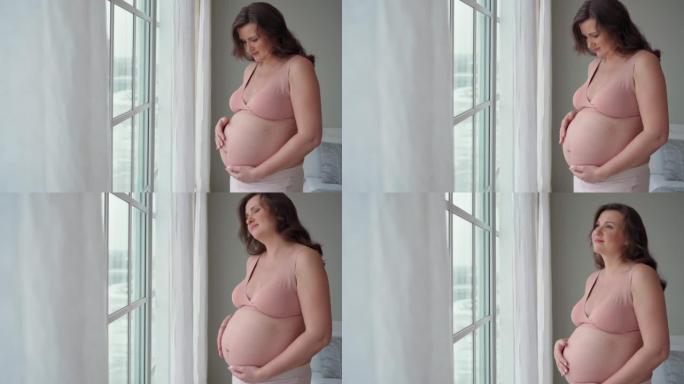 一个年轻漂亮的孕妇站在窗前抚摸她怀孕的腹部特写。期待孩子，做母亲，幸福，怀孕，