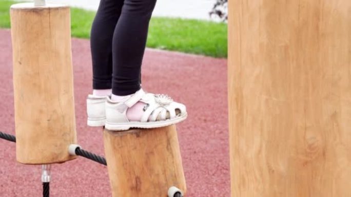 一个小女孩的脚在障碍赛绳梯上的特写镜头，上面装有原木进行平衡训练，孩子小心翼翼地走过去。木制现代儿童
