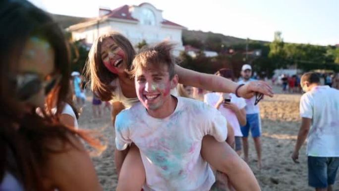 微笑的男人背着漂亮女人，在海滩的胡里节上都沾满了彩色粉末。在印度教的色彩，爱，团结的节日中，夫妻与朋