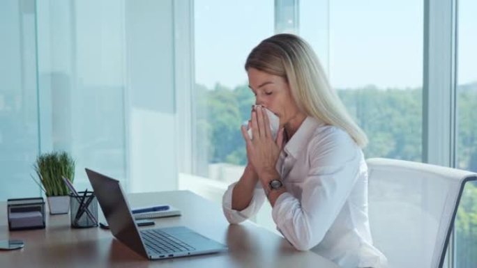 咳嗽，打喷嚏，流鼻涕。坐在工作场所的女商人在笔记本电脑上咳嗽，并用全景窗户的餐巾纸擦拭鼻子。感冒，流