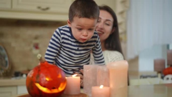 快乐的小男孩吹蜡烛，模糊的爱的女人在后台微笑着说话。快乐的高加索儿子的肖像与母亲在家里的厨房里享受万