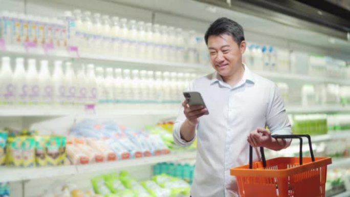 亚洲男性购物者在超市里推着购物车，看着智能手机，浏览智能手机，搜索商品或商品清单