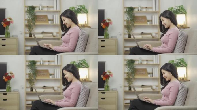 亚洲女人坐在沙发上，膝上放着笔记本电脑，打字而不是表现出快乐的表情，兴奋的女人看着笔记本电脑上的网上