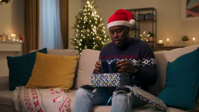 平安夜。一个非洲裔男人戴着圣诞老人的帽子坐在沙发上，打开礼物。他打开盒子，看到不需要的温暖耳机。男孩