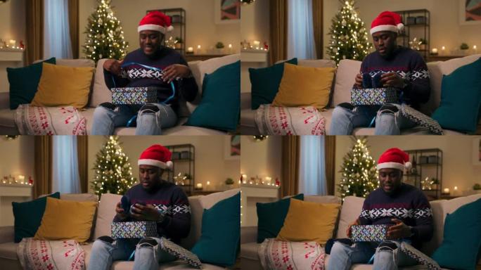 平安夜。一个非洲裔男人戴着圣诞老人的帽子坐在沙发上，打开礼物。他打开盒子，看到不需要的温暖耳机。男孩