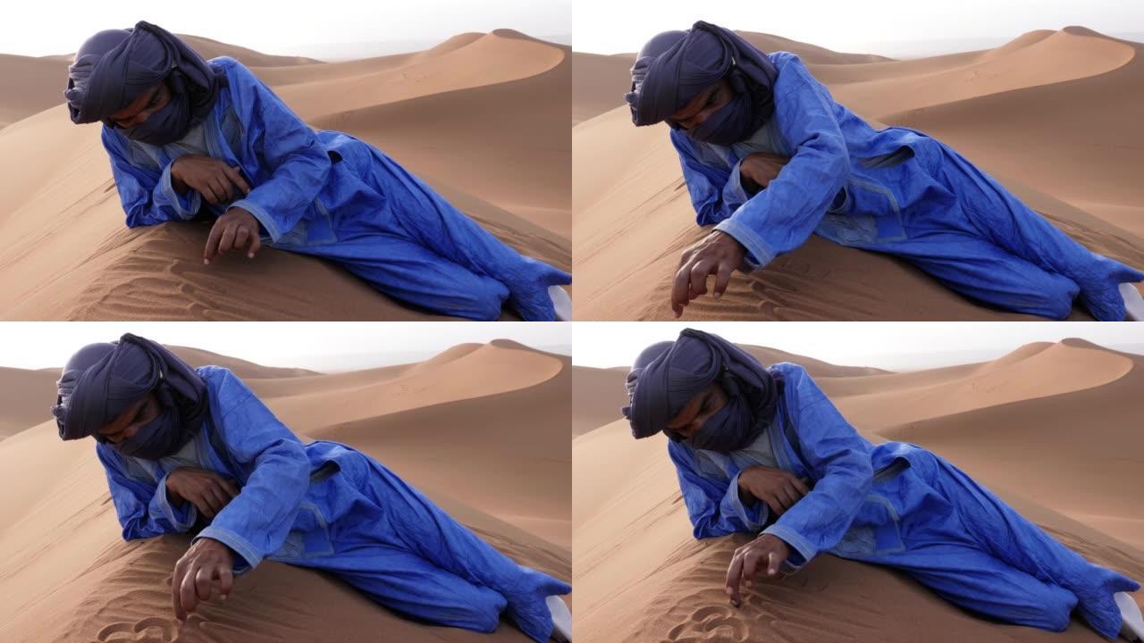 一名摩洛哥男子穿着蓝色的gandoura，djellaba和头巾，躺在摩洛哥Chigaga沙漠的沙丘