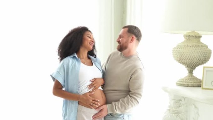 白人摸着他怀孕的黑人妻子的肚子。孕妇和慈爱的丈夫在家里拥抱肚子。多民族夫妇在怀孕的腹部感受婴儿的运动