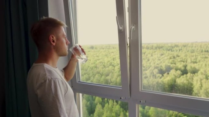 一个穿着便服的年轻人一边喝着干净的水，一边从窗户欣赏公园的景色