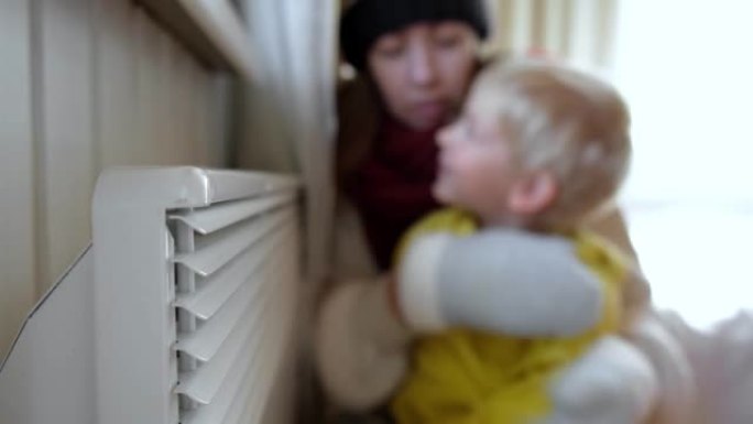 冷冻的母亲和儿子穿着毛衣摇晃和冷冻，冬天的寒冷拥抱在电加热器上。女孩和孩子正在暖气片附近暖手。呆在家