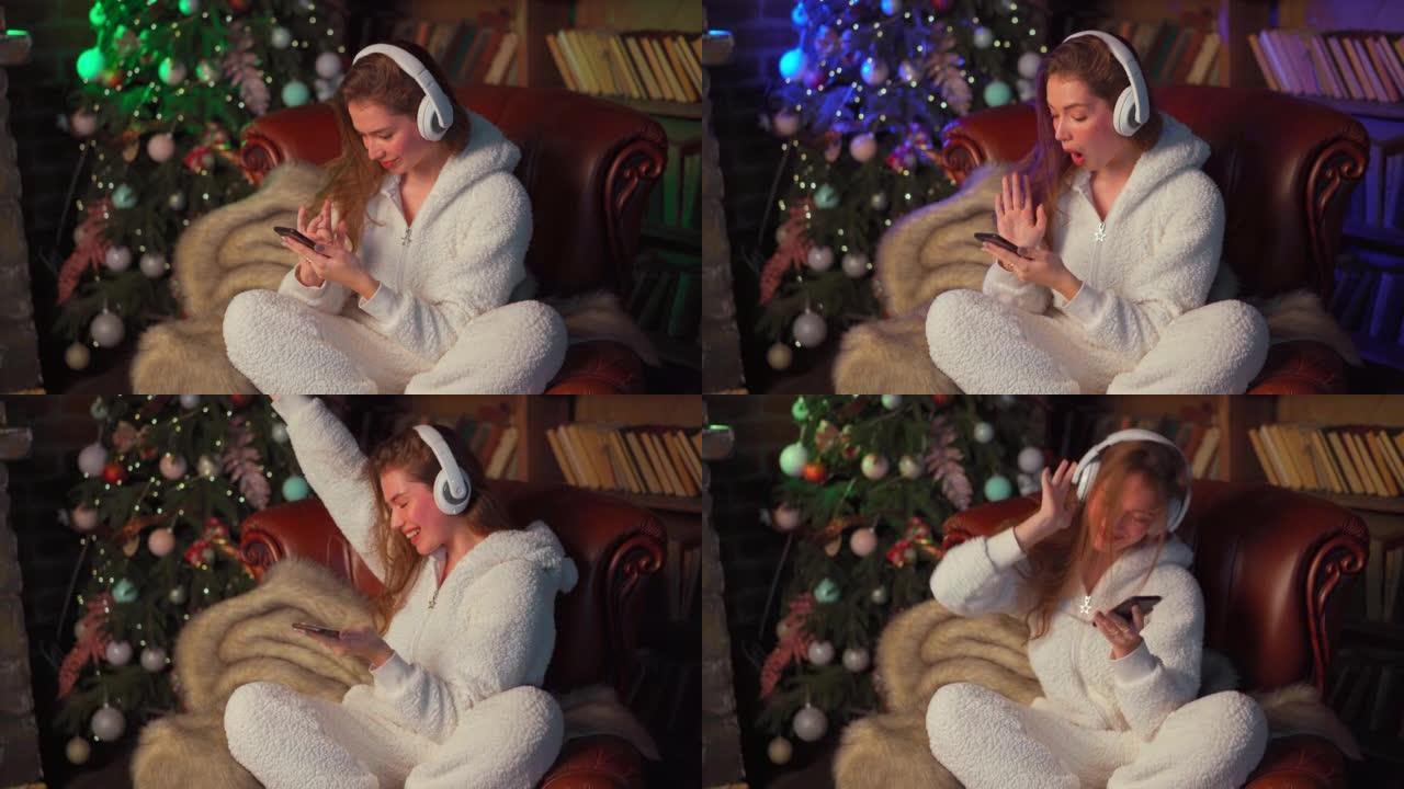 年轻女子耳机坐在圣诞树附近的皮革扶手椅上手持智能手机选择一首歌