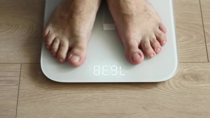 男子称自己 -- 男性赤脚在家踩着白色数字地磅: 特写镜头。测量体重，控制，健康和饮食概念