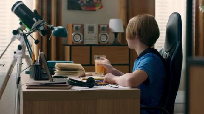 微笑男孩的肖像坐在笔记本电脑前的桌子上的椅子上，学生在线上课，听老师的话，做笔记，在家勤奋学习