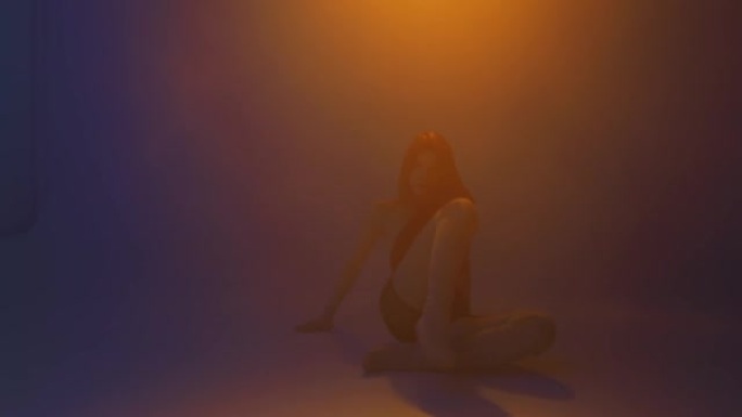 性感的女孩在彩色的灯光下跳舞在烟雾缭绕的工作室。热情的时尚舞蹈在烟雾中。在舞池中表演的女舞者