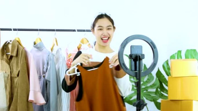 年轻的亚洲女性的4k视频直播显示衣服在智能手机摄像头前聊天并在家里的onling商店出售。
