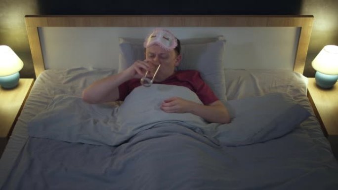 失眠男子晚上在床上喝安眠药，然后用玻璃杯中的水入睡，用睡眠面罩遮住眼睛，俯视图