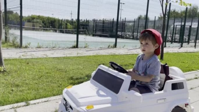 一个小男孩坐在白色玩具汽车上，在新鲜的空气中骑在绿色的公园里
