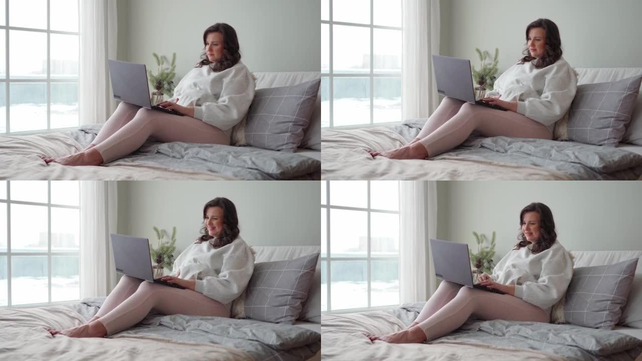 年轻漂亮的孕妇穿着舒适的衣服坐在床上，看着笔记本电脑上的东西。未来孕产的概念