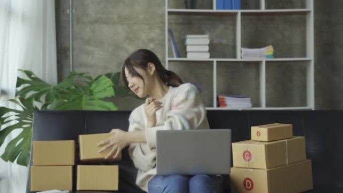 亚洲妇女坐在沙发上，坐在家里的盒子和笔记本电脑从客户网上业务中小企业销售概念