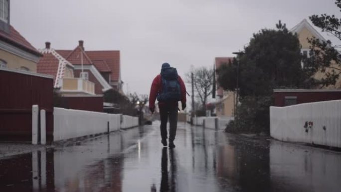 穿着冬装的背包的男人在雨中走在潮湿的柏油路上