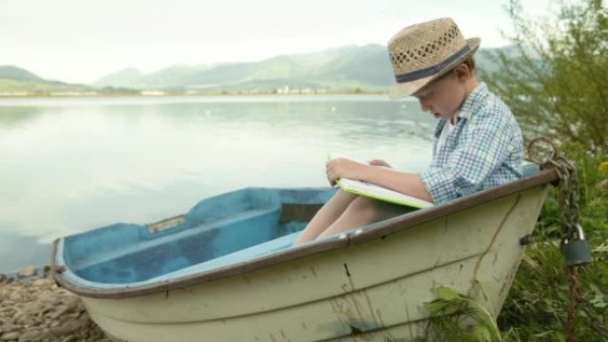 戴着草帽的小男孩坐在湖岸的乡村船上看书