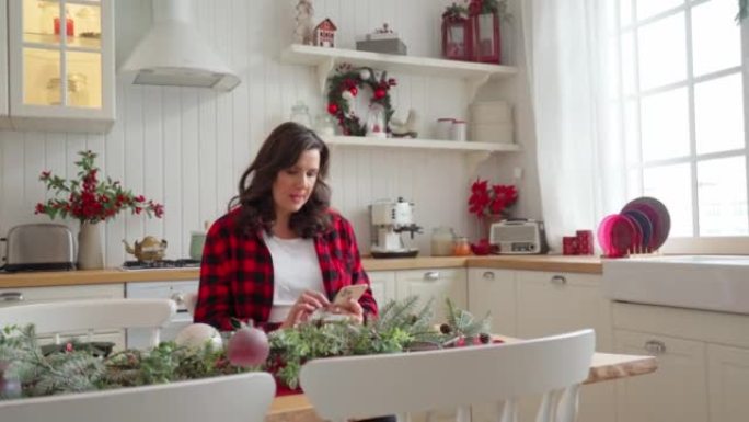 一名孕妇坐在明亮的大厨房的桌子旁，该厨房为圣诞节装饰，看着智能手机中的东西。订餐、网上购物、交流