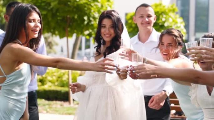 新娘喝香槟，与兴奋的朋友碰杯，欢呼。穿着相同礼服的伴郎和漂亮的伴娘在公园的婚礼上玩得很开心。慢动作。
