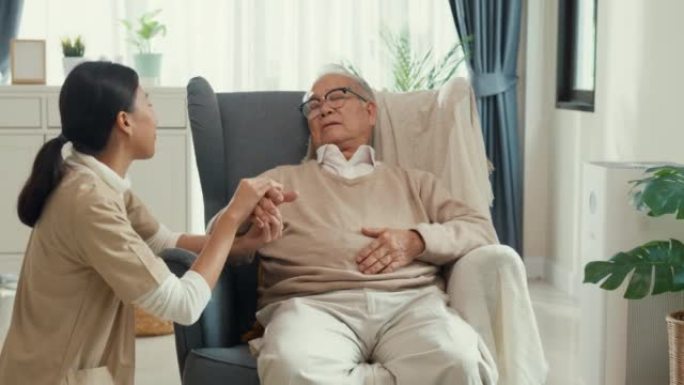 年轻的亚洲女性护理人员手持老年男性的手在家中的翼椅或扶手椅上照顾老年患者。