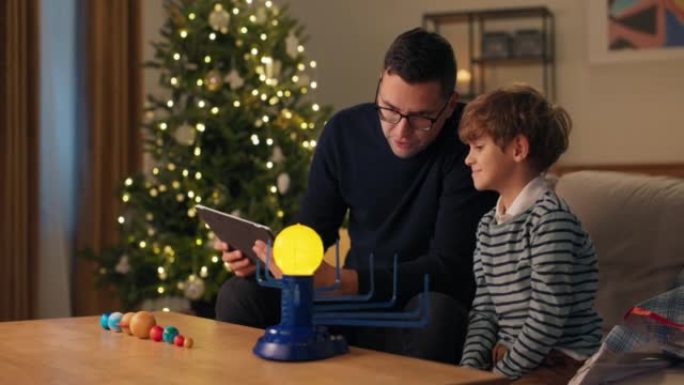 父亲在圣诞节晚上教孩子在平板电脑上查看玩具说明，发光的太阳系模型，在适当的地方安排行星