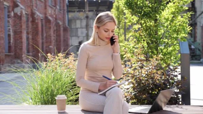 年轻的金发女孩在外面用手机说话时在笔记本上做笔记。手提电脑的美女在网上进行远程远程工作，坐在大学附近