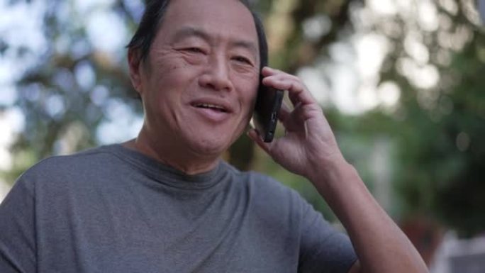 快乐的亚洲高级男子在城市街道上行走时打电话。一个中年老人与手机设备交谈的肖像