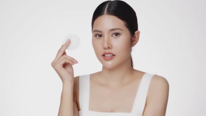 迷人美丽的年轻亚洲妇女清洁脸与棉垫隔离在白色背景。