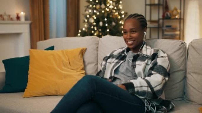 穿着方格衬衫的非洲外表迷人的女孩坐在客厅的沙发上。沙发后面闪烁着圣诞树。女孩戴着耳机听音乐，跟着唱歌