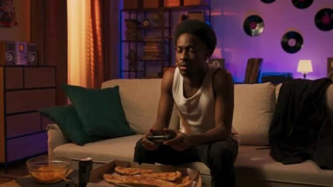一个非洲裔美国人后裔的人伸手去拿控制台上的垫子，在房间里度过时光，舒适地坐在沙发上吃外卖披萨，在电视