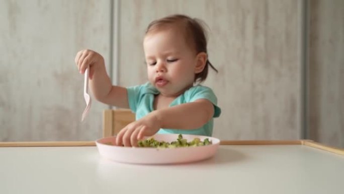一个女孩小白种人的孩子蹒跚学步的孩子坐在家里的桌子上独自吃食物，使用塑料叉子，童年成长和发展的理念拷