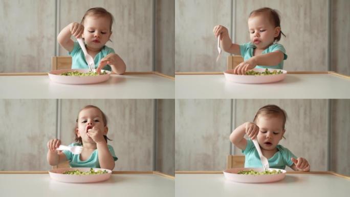 一个女孩小白种人的孩子蹒跚学步的孩子坐在家里的桌子上独自吃食物，使用塑料叉子，童年成长和发展的理念拷