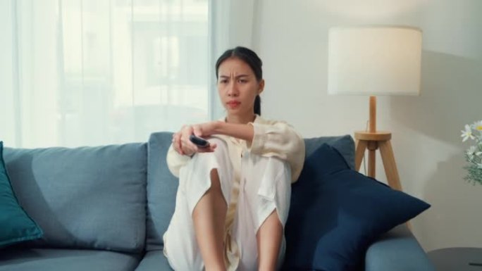 年轻的亚洲无聊的女人在沙发上点击遥控器更改兴趣频道电影或在电视上播放在线视频点播，在家点播视频。