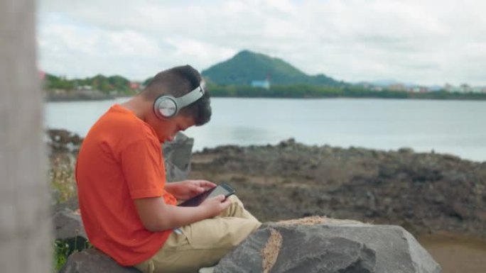 坐在海边看数字平板电脑和听音乐的孩子，中等镜头