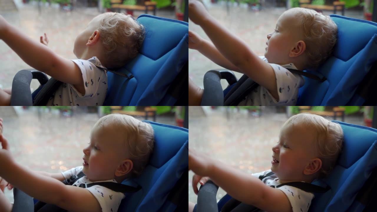 婴儿推车上的孩子看着一场暴雨坐在夏季咖啡馆的树冠下，婴儿的脸顶着雨滴。