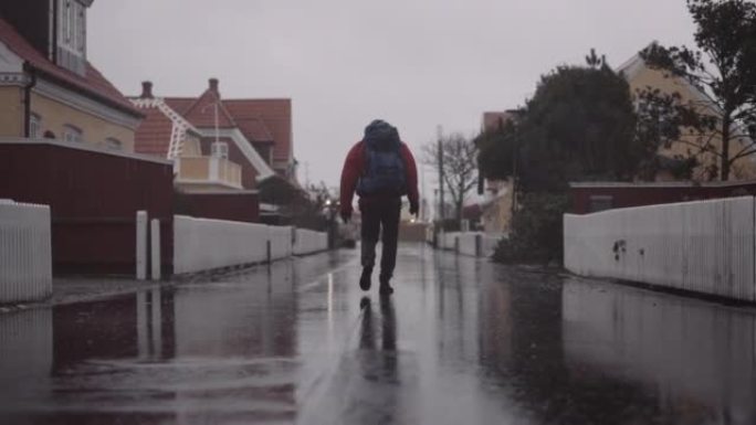 穿着冬装的背包的男人在雨中走在潮湿的柏油路上