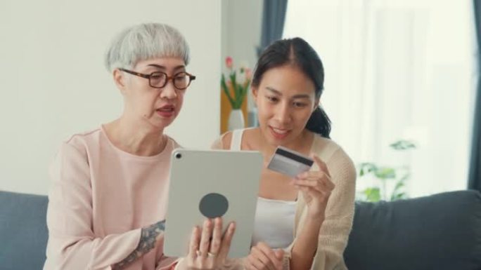 快乐的亚洲家庭，年轻的女儿和年长的妈妈教年长的妈妈使用数字平板电脑在线购物使用信用卡支付在线应用在家