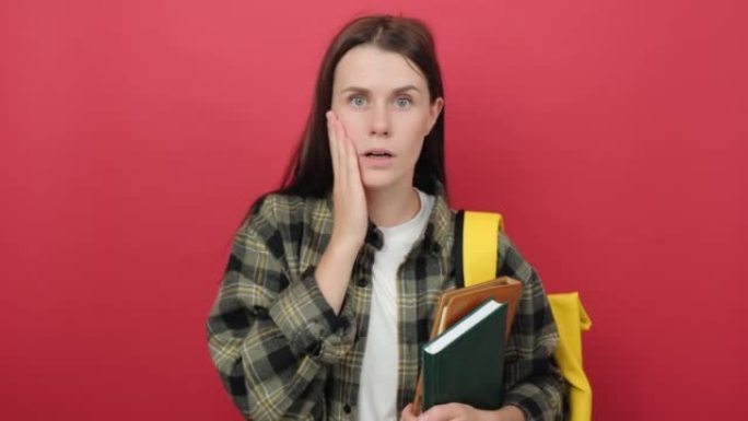 穿着衬衫和黄色背包的震惊的年轻学生女子用手捂着书的嘴，孤立在工作室的红色背景墙上。高中教育大学学院理