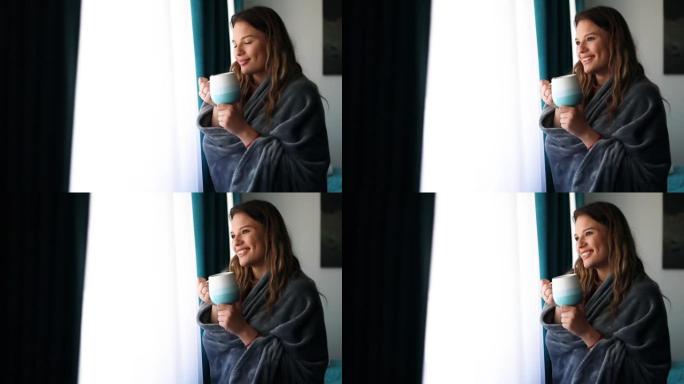 美丽的年轻女子披着毯子站在窗户后面喝茶