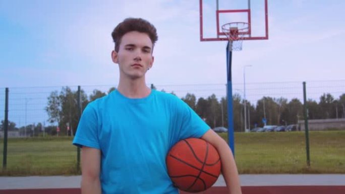严肃的少年带着篮球在夏日日落时看着相机，电影般的