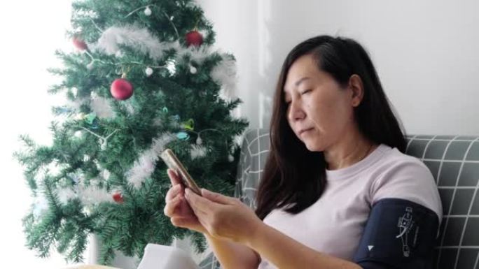 亚洲成年女性坐在家里圣诞树附近的沙发上，与医生交谈，进行血压测试，生活方式概念。