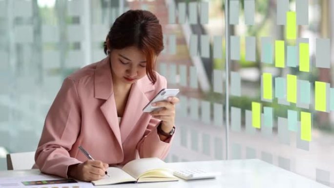 亚洲女商人在记事本上写笔记，在她的智能手机上计划她每月的工作日程。