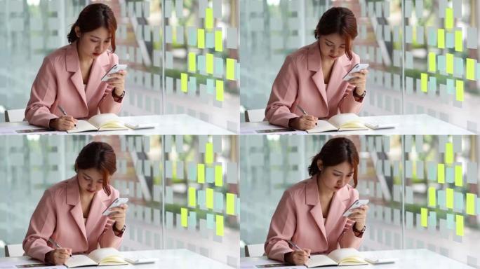 亚洲女商人在记事本上写笔记，在她的智能手机上计划她每月的工作日程。