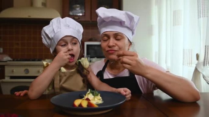 穿着白色厨师帽和围裙的高加索可爱的小女孩，和妈妈在家庭厨房里吃美味的冰淇淋