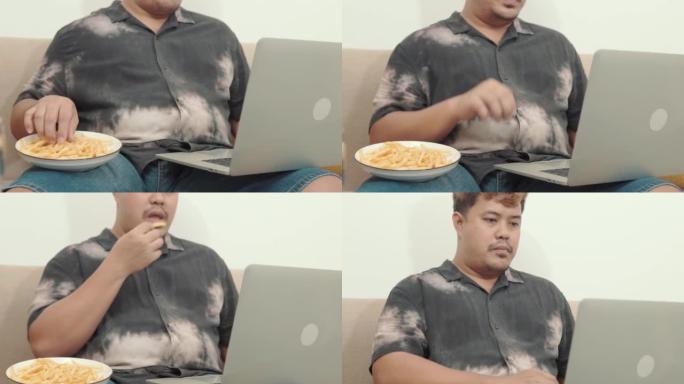 4k视频镜头的大身材男人吃炸薯条和在家里的沙发上使用笔记本电脑，不健康的生活方式概念。