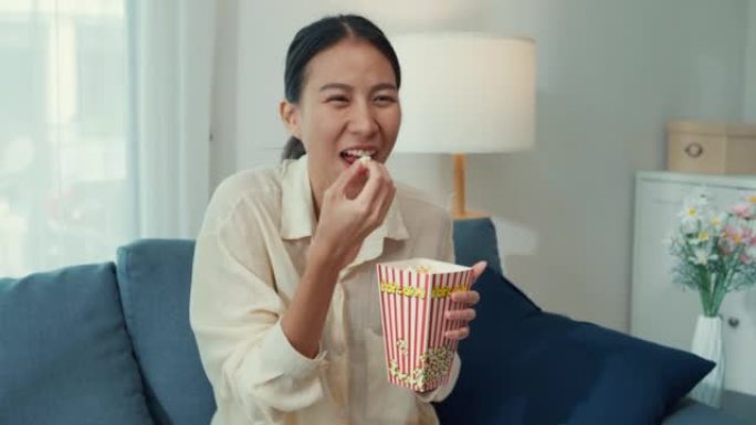 年轻的亚洲女孩在沙发上吃爆米花有趣的笑声观看喜剧频道电影或情景喜剧在电视上播放在线视频点播，在家点播