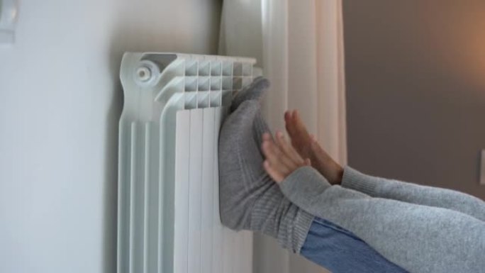 女性把脚放在暖气散热器上的灰色袜子里试图在寒冷的公寓里热身